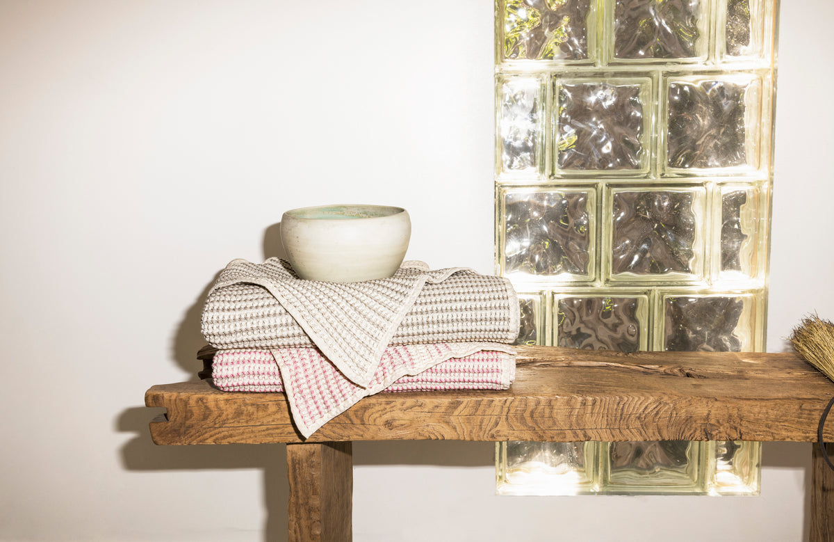 Stacked Sand and Shell baby receiving towels on a wooden bench, with a cream ceramic bowl, showing a natural material still life.