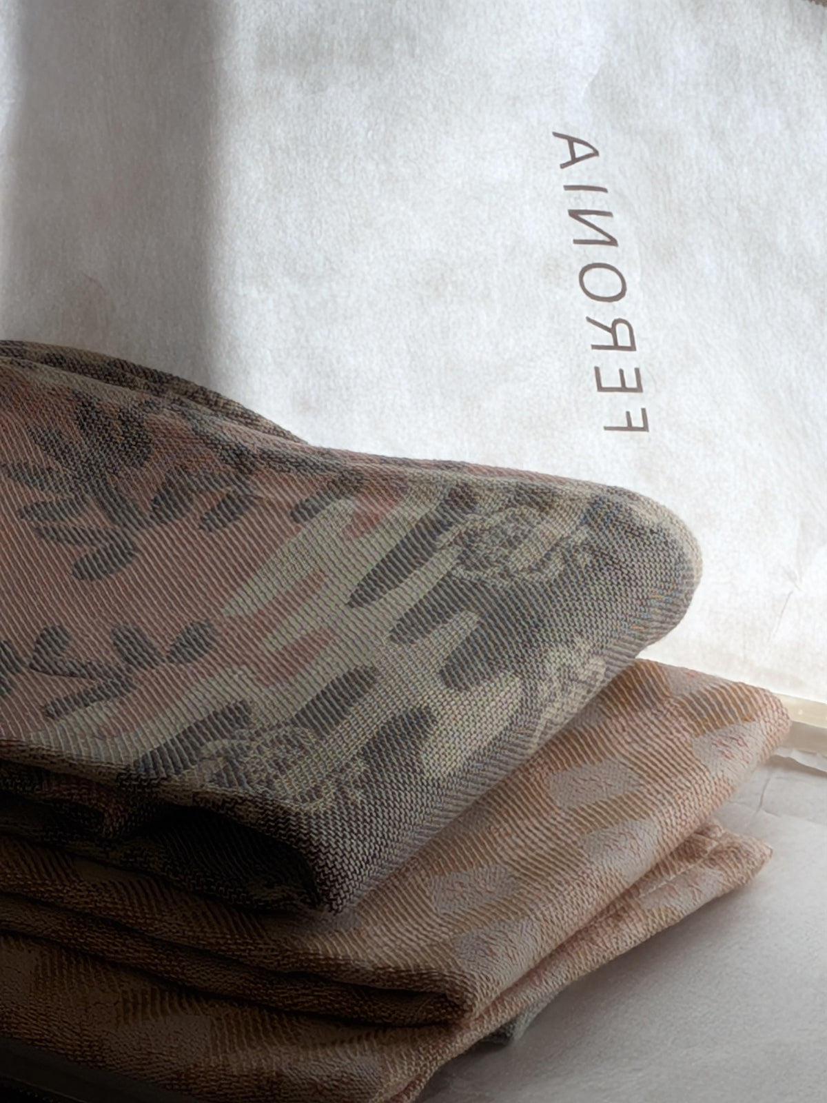 Sesame/Plum jacquard kitchen towels stacked in soft light beside the Feronia gift pouch